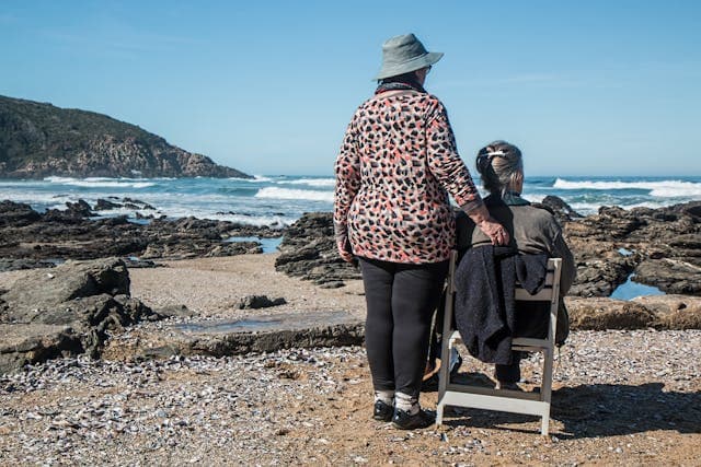 resident sat in chair on beach with carer stood alongside